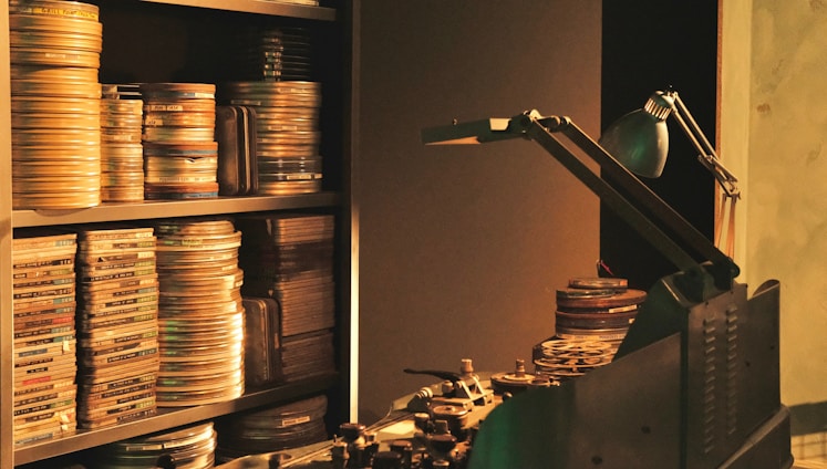 Film reels are stacked on shelves and a table, alongside a vintage film editing machine. A desk lamp provides dim, focused light over the area.