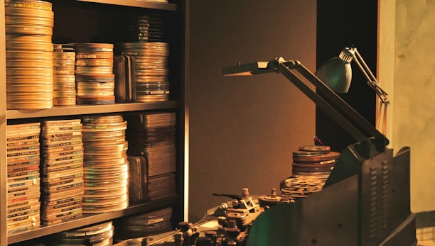 Film reels are stacked on shelves and a table, alongside a vintage film editing machine. A desk lamp provides dim, focused light over the area.