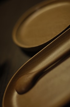 A close-up view of wooden kitchenware, including a spoon and a tray. The items are carved elegantly, showcasing smooth textures and a natural brown hue. The shallow depth of field creates a soft blur effect in the background.