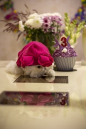 A small dog wearing a bright pink hat rests its head on a reflective surface. Beside the dog is a purple cupcake with a number three candle on top, suggesting a celebration or birthday. In the background, there is a bouquet of mixed flowers in various colors, including white and purple.