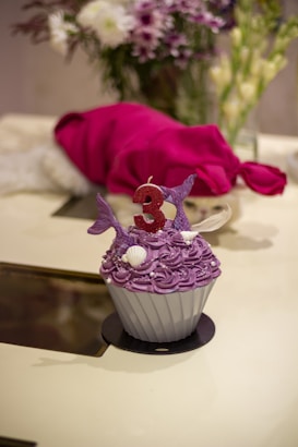 A purple cupcake with elaborate swirling frosting, decorated with mermaid tails and seashells, and topped with a red candle in the shape of the number three. In the background, there is a blurred bouquet of flowers and a red fabric.