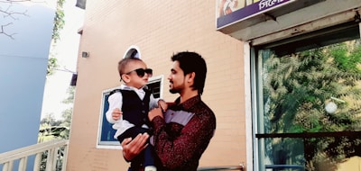 A man holds a child dressed in formal attire with sunglasses on, standing outside a building. The man is gazing at the child affectionately. The background includes a brick wall, a window showing greenery, and a sign partially visible above.
