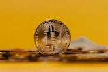 A shiny gold Bitcoin coin is prominently displayed against a bright yellow background. The coin's design includes detailed engravings, with the recognizable Bitcoin symbol at the center. Other coins appear slightly out of focus in the foreground, emphasizing the main Bitcoin.