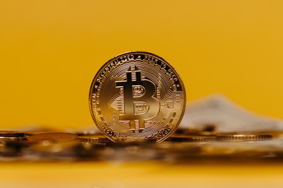 A shiny gold Bitcoin coin is prominently displayed against a bright yellow background. The coin's design includes detailed engravings, with the recognizable Bitcoin symbol at the center. Other coins appear slightly out of focus in the foreground, emphasizing the main Bitcoin.
