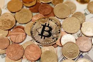 A large assortment of coins is scattered over paper currency, with a prominent Bitcoin coin at the center symbolizing cryptocurrency.