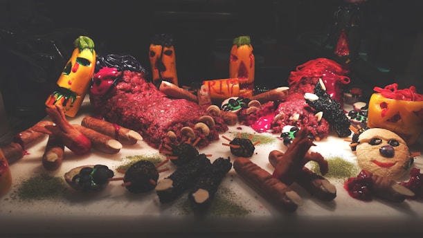 A Halloween-themed food display featuring various dishes creatively shaped to resemble eerie and spooky figures. The scene includes carved vegetables and meats dyed in deep reds and greens, resembling monsters or ghouls. The layout is dramatic, with props like dismembered fingers and RIP tombstones, creating a spooky atmosphere.
