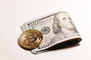 A stack of US hundred-dollar bills is folded and placed on a light background. Beside it, there is a shiny gold coin featuring the Bitcoin symbol. The image juxtaposes traditional currency with cryptocurrency.