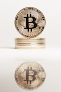 A stack of golden coins with the Bitcoin symbol prominently displayed on the top coin. The image is well-lit and sharp, featuring a clear reflection of the coin stack on a reflective surface below.