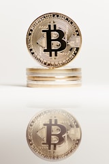 A stack of golden coins with the Bitcoin symbol prominently displayed on the top coin. The image is well-lit and sharp, featuring a clear reflection of the coin stack on a reflective surface below.