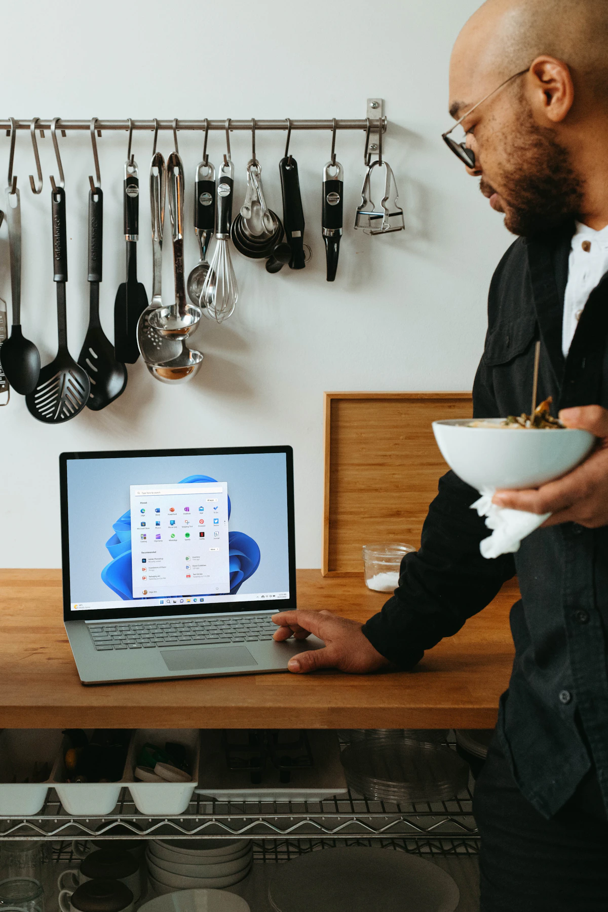 A man touching a Windows laptop while holding a bowl of food in a kitchen.