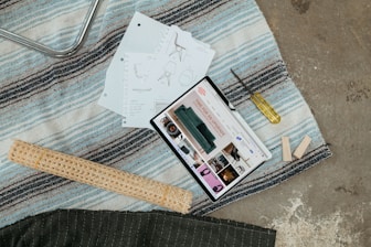 A scene with a tablet displaying interior design content, placed on a striped rug alongside design sketches, a screwdriver, wooden planks, and a rolled mat.