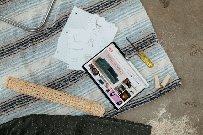 A scene with a tablet displaying interior design content, placed on a striped rug alongside design sketches, a screwdriver, wooden planks, and a rolled mat.