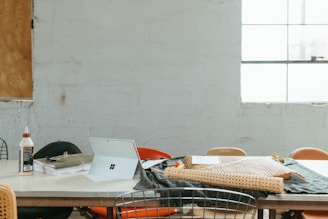 A workspace table with various items including a bottle of glue, a stack of papers bound with string, a tablet with a stand, a piece of woven material, and fabric. The setting is minimalistic with a large window in the background and chairs around the table.