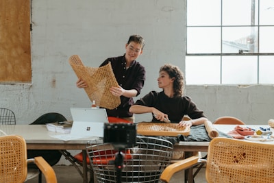 Two people are engaged in a craft activity, working with woven materials at a table. The setting appears to be a bright workspace or studio with natural light coming through a large window. Various tools and materials are spread across the table, and there is a laptop visible, suggesting a blend of traditional craft and modern technology.