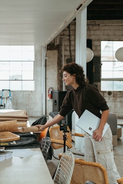 A person with curly hair holding a laptop stands near a table in a spacious, industrial-style room with large windows. The room features various items such as chairs, fabric materials, and cameras. The exposed brick walls and high ceiling contribute to a creative, workshop atmosphere.