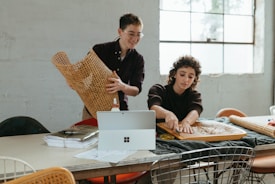 Two people are engaged in a craft activity at a table. One person is holding a piece of woven material, while the other is working on a similar piece placed on the table. A laptop, papers, and tools are also present on the table. The setting is a brightly lit room with large windows.