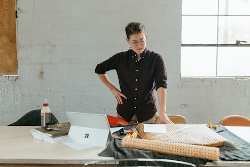 A person stands at a table covered with fabric rolls and a laptop. They seem focused and are holding a piece of paper, possibly involved in design or planning. The setting is a bright room with large windows and a minimalist, industrial feel.