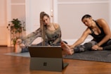 Two people are engaged in a stretching exercise on yoga mats in a cozy room with wooden flooring. A tablet or laptop is placed on the floor in front of them, possibly for following a virtual class. The room is decorated with potted plants and has a warm, inviting atmosphere.