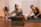 Two people are engaged in a stretching exercise on yoga mats in a cozy room with wooden flooring. A tablet or laptop is placed on the floor in front of them, possibly for following a virtual class. The room is decorated with potted plants and has a warm, inviting atmosphere.