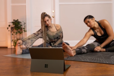 Two people are engaged in a stretching exercise on yoga mats in a cozy room with wooden flooring. A tablet or laptop is placed on the floor in front of them, possibly for following a virtual class. The room is decorated with potted plants and has a warm, inviting atmosphere.