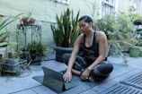 A woman sits cross-legged on a mat outdoors, surrounded by various potted plants. She is focused on a tablet or laptop resting on the mat in front of her. The area is lush with greenery, and the wall behind is painted in a muted color. Her attire is camouflage patterned, and she appears to be engaged in an activity such as yoga, meditation, or work.