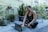 A woman sits cross-legged on a mat outdoors, surrounded by various potted plants. She is focused on a tablet or laptop resting on the mat in front of her. The area is lush with greenery, and the wall behind is painted in a muted color. Her attire is camouflage patterned, and she appears to be engaged in an activity such as yoga, meditation, or work.
