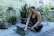 A woman sits cross-legged on a mat outdoors, surrounded by various potted plants. She is focused on a tablet or laptop resting on the mat in front of her. The area is lush with greenery, and the wall behind is painted in a muted color. Her attire is camouflage patterned, and she appears to be engaged in an activity such as yoga, meditation, or work.