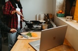 A person wearing a red flannel shirt is cooking on a stove with a wok. A chopping board on the counter has diced vegetables and a knife. A laptop is open on the counter, and there are various kitchen items nearby, including a bowl of oranges on the windowsill.