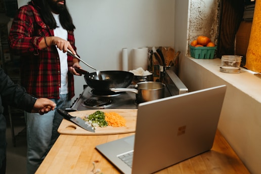 A person wearing a red flannel shirt is cooking on a stove with a wok. A chopping board on the counter has diced vegetables and a knife. A laptop is open on the counter, and there are various kitchen items nearby, including a bowl of oranges on the windowsill.