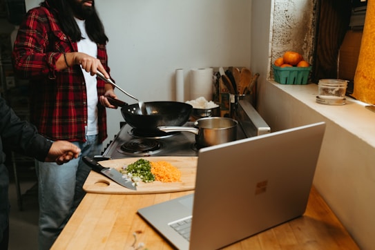 A person wearing a red flannel shirt is cooking on a stove with a wok. A chopping board on the counter has diced vegetables and a knife. A laptop is open on the counter, and there are various kitchen items nearby, including a bowl of oranges on the windowsill.