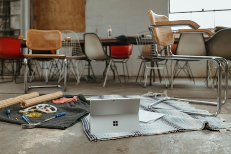 A workspace with various chairs around a long table alongside a cluttered area on the floor. An open laptop sits on top of a striped rug surrounded by tools like pliers, screwdrivers, and wooden sticks, indicating a DIY or creative project setting.
