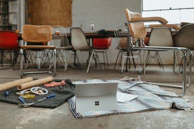A workspace with various chairs around a long table alongside a cluttered area on the floor. An open laptop sits on top of a striped rug surrounded by tools like pliers, screwdrivers, and wooden sticks, indicating a DIY or creative project setting.