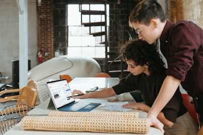 Two people are collaborating over a laptop and blueprint on a table in an industrial-style room with brick walls and metal chairs. One person is seated, pointing at the blueprint, while the other is standing, looking at the laptop screen. A fire extinguisher and some rolled-up materials are visible in the background.