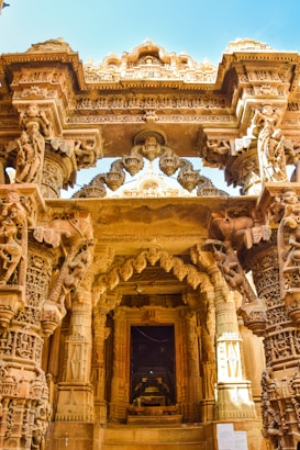 Intricate stone carvings and sculptures adorn a beautifully designed entrance of an ancient temple. The architecture features detailed pillars and archways with ornate patterns, depicting figures and mythological motifs.