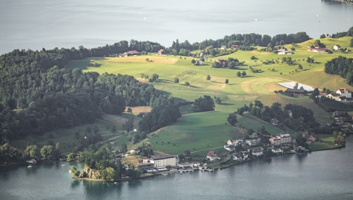 A scenic aerial view of a rural area featuring lush green fields and dense forests. Multiple houses dot the landscape, with a larger building near the water's edge. The serene body of water in the foreground reflects the surrounding environment.