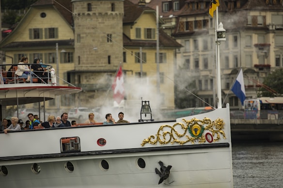 A historic steamship with ornate gold detailing on its bow is sailing on a river or lake, with a backdrop of European-style buildings and a Swiss flag visible amidst steam or smoke.