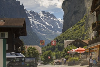 A picturesque village scene with traditional wooden chalets flanking a street. Swiss flags hang prominently, and there are people walking along the street. In the background, towering snow-capped mountains rise sharply, partially covered in shadow. A waterfall cascades down a steep cliff, adding to the serene mountain landscape.