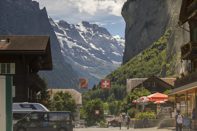 A picturesque village scene with traditional wooden chalets flanking a street. Swiss flags hang prominently, and there are people walking along the street. In the background, towering snow-capped mountains rise sharply, partially covered in shadow. A waterfall cascades down a steep cliff, adding to the serene mountain landscape.