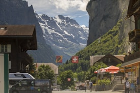 A picturesque village scene with traditional wooden chalets flanking a street. Swiss flags hang prominently, and there are people walking along the street. In the background, towering snow-capped mountains rise sharply, partially covered in shadow. A waterfall cascades down a steep cliff, adding to the serene mountain landscape.