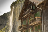 A traditional wooden chalet with green window shutters and flower boxes on the balconies stands prominently in the foreground. In the background, a waterfall cascades down a steep, rocky cliff surrounded by lush green trees.