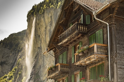 A traditional wooden chalet with green window shutters and flower boxes on the balconies stands prominently in the foreground. In the background, a waterfall cascades down a steep, rocky cliff surrounded by lush green trees.