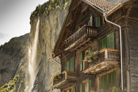 A traditional wooden chalet with green window shutters and flower boxes on the balconies stands prominently in the foreground. In the background, a waterfall cascades down a steep, rocky cliff surrounded by lush green trees.