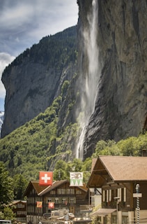 A picturesque mountainous landscape features a tall waterfall cascading down a steep rock face amidst green foliage. Below, a quaint village with traditional alpine architecture is adorned with Swiss flags, lending a sense of national pride. The scene captures both the grandeur of nature and the charm of rural life under a clear blue sky.