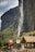 A picturesque mountainous landscape features a tall waterfall cascading down a steep rock face amidst green foliage. Below, a quaint village with traditional alpine architecture is adorned with Swiss flags, lending a sense of national pride. The scene captures both the grandeur of nature and the charm of rural life under a clear blue sky.