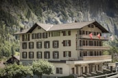 A multi-story traditional building with white walls and brown shutters labeled 'Hotel Staubbach' stands against a backdrop of steep rocky cliffs and lush green trees. Several balconies adorned with flags offer views of the picturesque surroundings.
