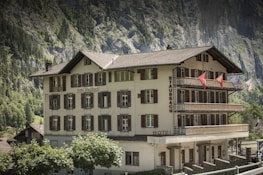 A multi-story traditional building with white walls and brown shutters labeled 'Hotel Staubbach' stands against a backdrop of steep rocky cliffs and lush green trees. Several balconies adorned with flags offer views of the picturesque surroundings.