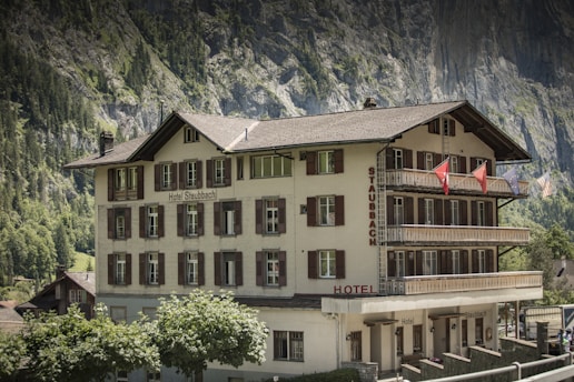 A multi-story traditional building with white walls and brown shutters labeled 'Hotel Staubbach' stands against a backdrop of steep rocky cliffs and lush green trees. Several balconies adorned with flags offer views of the picturesque surroundings.