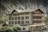 A multi-story traditional building with white walls and brown shutters labeled 'Hotel Staubbach' stands against a backdrop of steep rocky cliffs and lush green trees. Several balconies adorned with flags offer views of the picturesque surroundings.