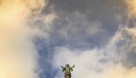 A statue of a saint with open arms stands prominently against a backdrop of a partly cloudy sky. The statue appears to be atop a pillar or column, and the clouds surrounding it are illuminated by a warm golden light.