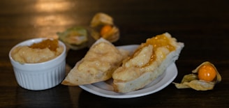 Two slices of bread with creamy spread and orange marmalade rest on a small white plate. Next to the plate is a ramekin containing the same spread and marmalade. In the background, there are a couple of physalis fruits, one with its husk partially open to reveal the bright orange fruit inside.